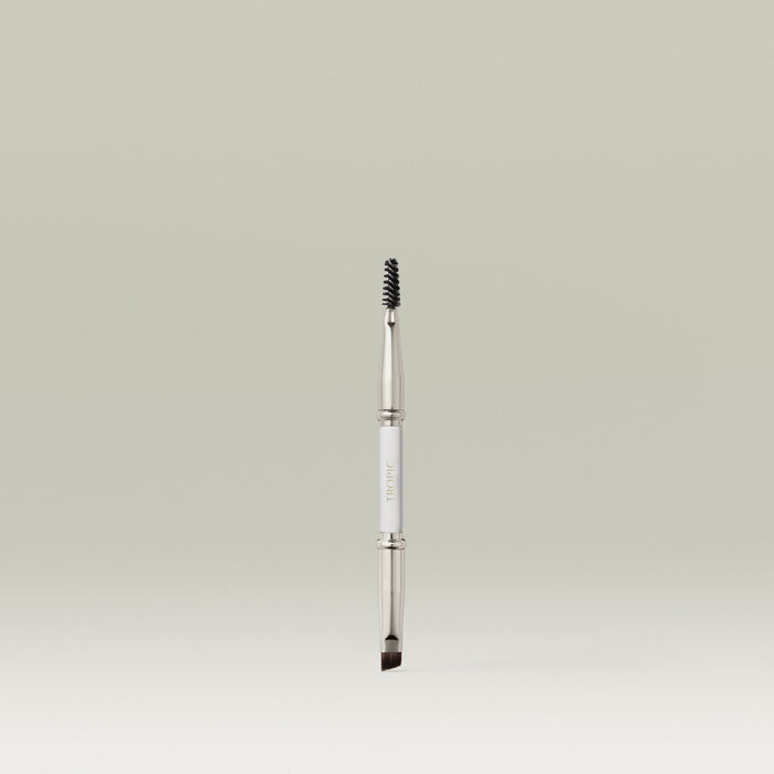 A dual-ended makeup brush featuring a spoolie on one end and an angled brush on the other, shown upright against a soft, neutral background.