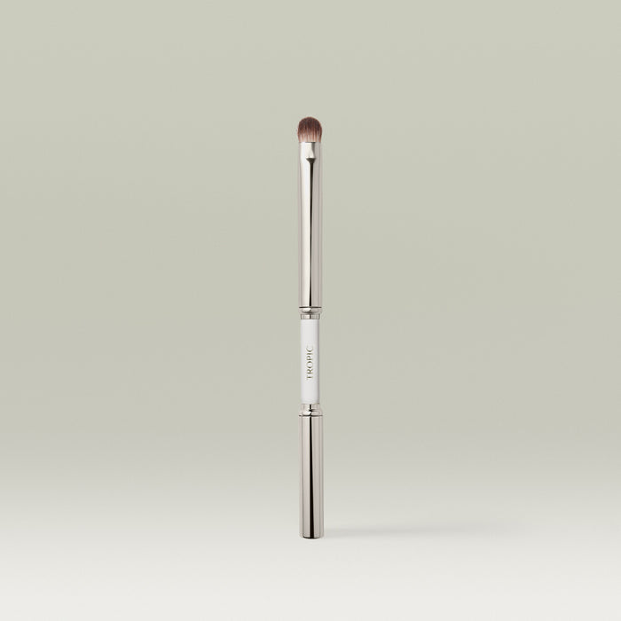 A sleek dual-ended metallic eye makeup brush shown upright with both ends capped, featuring a white center section printed with the brand name.