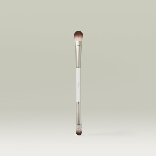 A dual-ended makeup brush with soft, rounded bristles and a sleek metallic finish, standing upright against a soft neutral background.