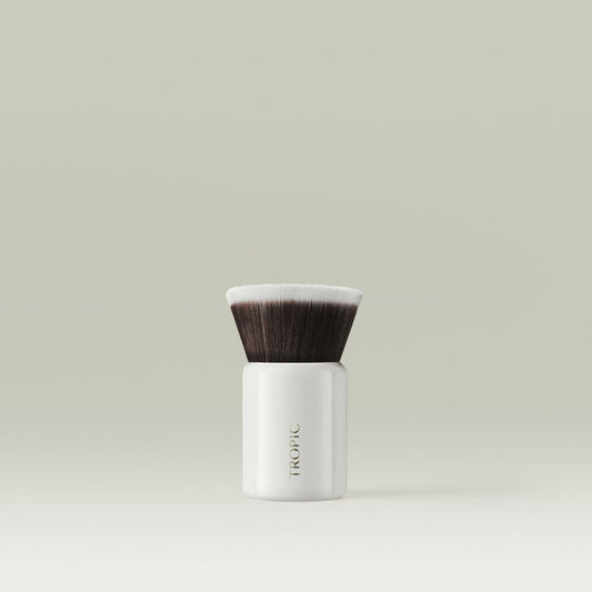 A compact kabuki makeup brush with dense, flat-topped bristles and a glossy white handle, standing upright against a soft, neutral background.