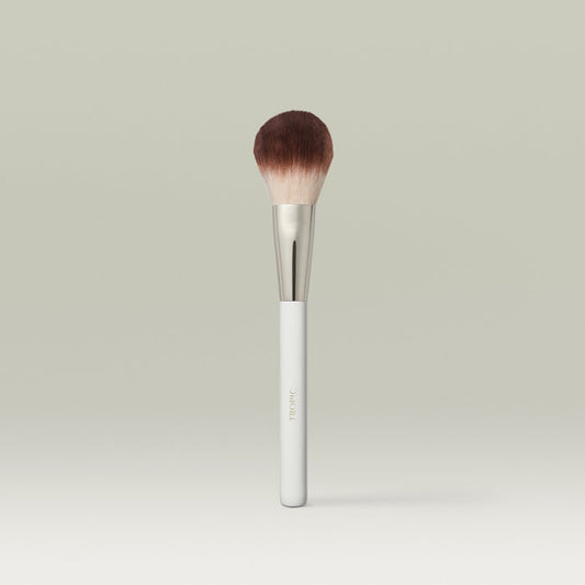 A fluffy, dome-shaped powder brush with soft brown bristles, a metallic silver ferrule, and a white handle, displayed upright against a soft, neutral background.