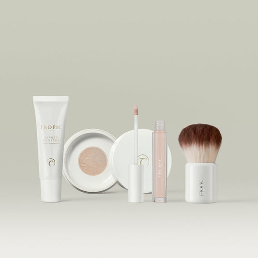 Tropic makeup collection in shade 1 displayed on a neutral background, featuring a white tube of Beauty Booster sheer foundation, an open compact with mesh applicator, a concealer with wand applicator, and a fluffy kabuki brush with a white base.