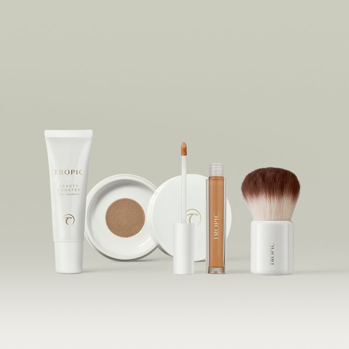 Tropic makeup collection in shade 7 displayed on a neutral background, featuring a white tube of Beauty Booster sheer foundation, an open compact with mesh applicator, a concealer with wand applicator, and a fluffy kabuki brush with a white base.