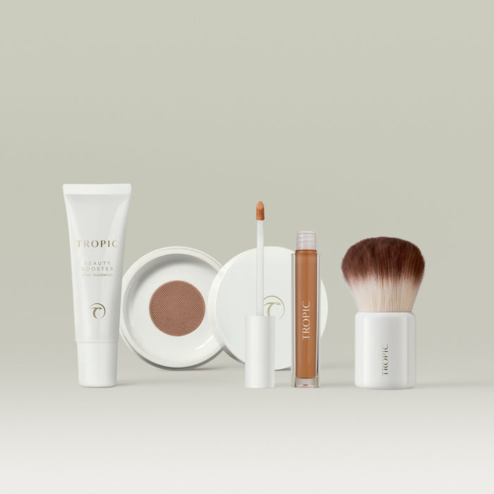 Tropic makeup collection in shade 8 displayed on a neutral background, featuring a white tube of Beauty Booster sheer foundation, an open compact with mesh applicator, a concealer with wand applicator, and a fluffy kabuki brush with a white base.