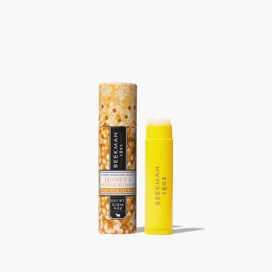Yellow uncapped tube of Beekman 1802's Honey & Orange Blossom Lip Balm, standing next to orange packaging tube that is designed with flowers, on a white background.