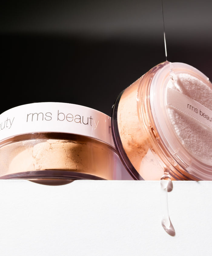 Close-up of two containers of Hydra Setting Powder by RMS Beauty with a drop of clear liquid dripping next to them, highlighting the clean beauty and natural ingredients.