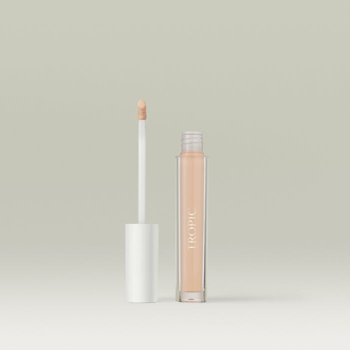 A  liquid concealer in a clear tube labelled “TROPIC,” shown open with its white-capped wand standing beside it against a soft, neutral background. In shade 2.
