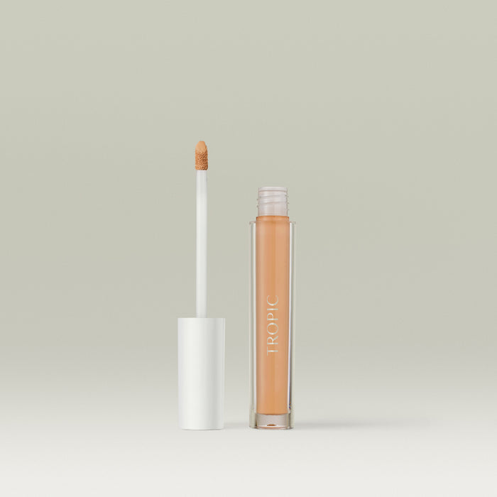 A  liquid concealer in a clear tube labelled “TROPIC,” shown open with its white-capped wand standing beside it against a soft, neutral background. In shade 4.