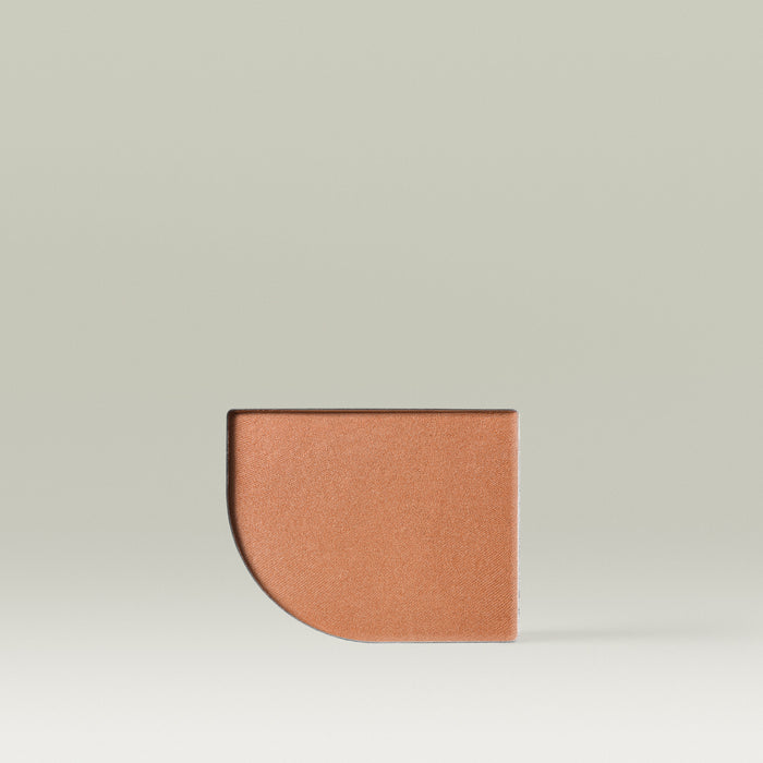 A Tropic Jet Set bronzer refill pan in the shade Havana Heat, shown as a warm, deep tan in a moon-shaped pressed powder pan against a soft neutral background.