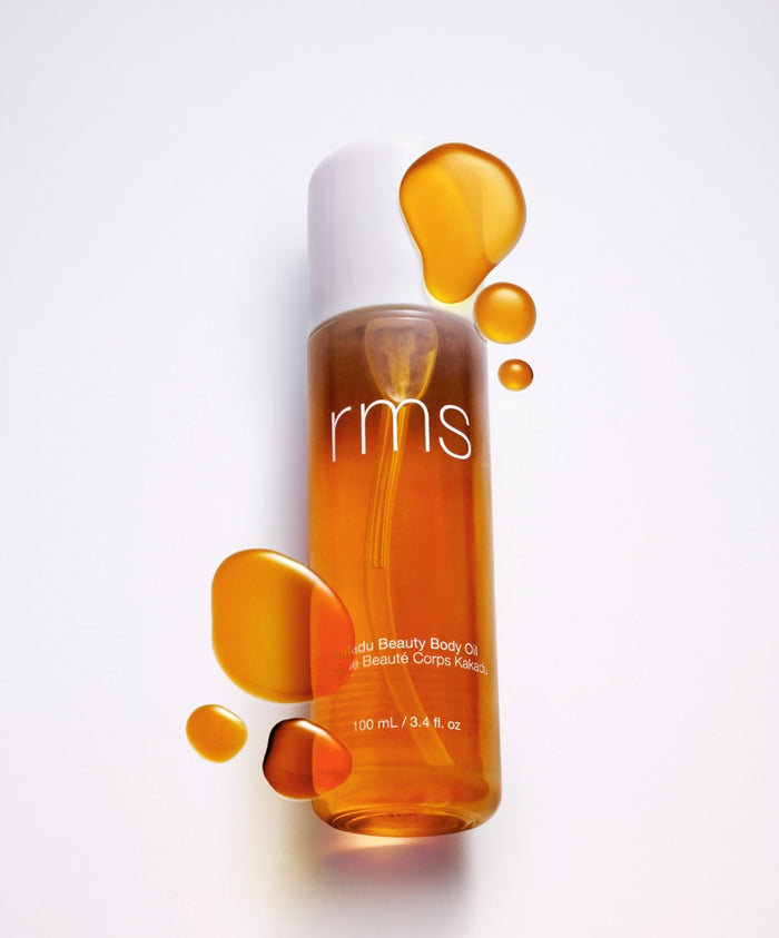 Kakadu Body Oil by RMS Beauty in a clear bottle with golden oil droplets around it, highlighting clean beauty, natural ingredients, and sustainable skincare.