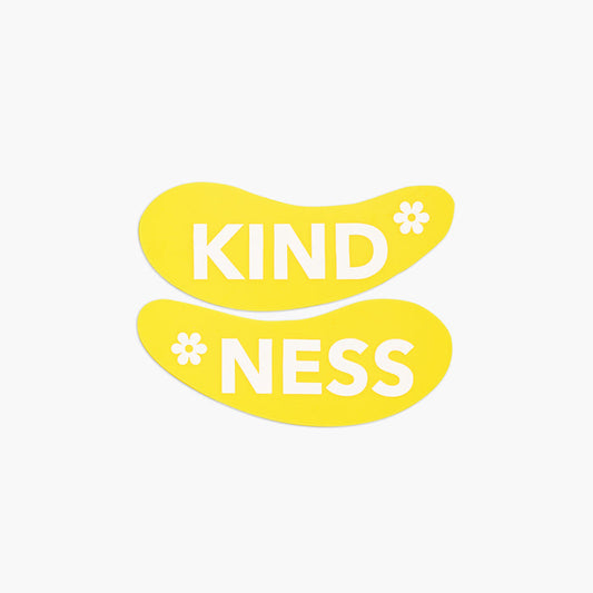 Beekman 1802's yellow Kindness Undereye Mask, with the left eye patch that spells "kind" on the top, and the right eye patch that spells "Ness" on the bottom and with a white flower on each eye patch.