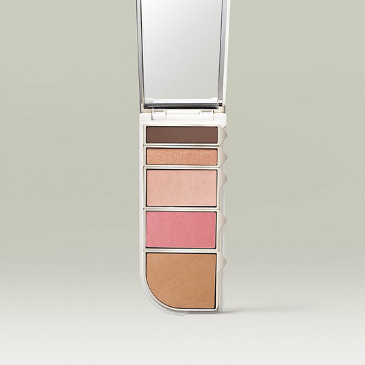 An open makeup palette displayed vertically, featuring five shades: a deep matte brown, a shimmery peach-gold, a soft champagne highlight, a bright pink blush, and a warm bronzer. A mirror is built into the lid above the pans.