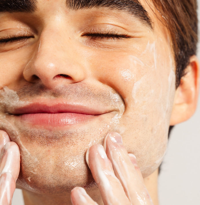 Up close shot of model's face while smiling with their eyes closed, and Beekman 1802's Vegan Goat Milk™ Oil Eliminating Foaming Gel Cleanser lathered on their face with their hands touching their chin.