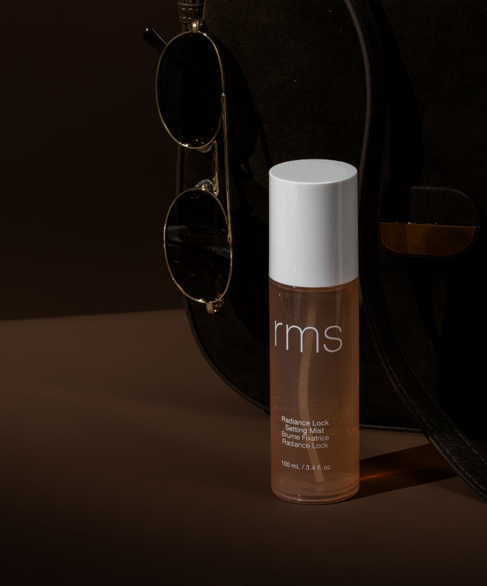 Radiance Lock Setting Mist bottle with a white cap placed beside a black handbag and gold-rimmed sunglasses, showcasing clean beauty and skincare infused makeup.