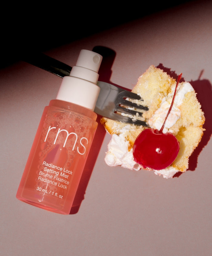 A bottle of Radiance Lock Setting Mist with a light peach-colored pump placed next to a fork holding a piece of cake topped with white cream and a bright red cherry on a soft pink surface.