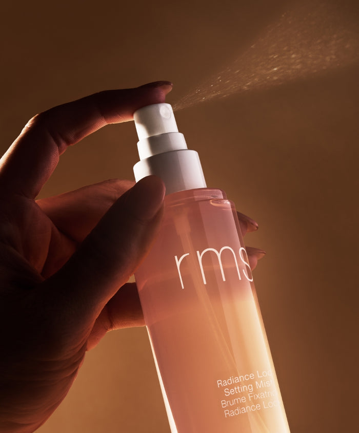 Hand pressing the spray nozzle of a pink translucent bottle labeled Radiance Lock Setting Mist, releasing a fine mist against a warm brown background.