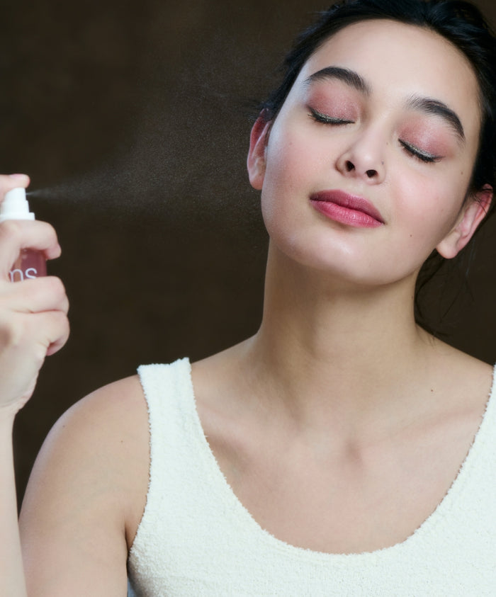 A woman with closed eyes gently sprays Radiance Lock Setting Mist on her face, showcasing clean beauty and natural ingredients in a calming skincare routine.