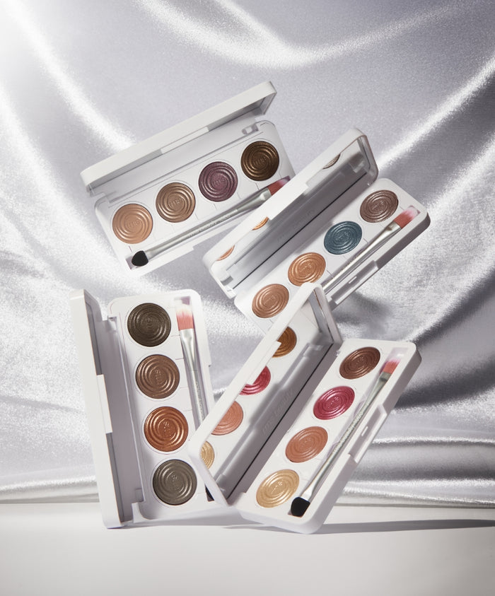 Four open ReDimension Hydra Eyes Quartet palettes showcasing a variety of creamy neutral and jewel-toned eyeshadows with applicator brushes on a shimmering silver fabric background.