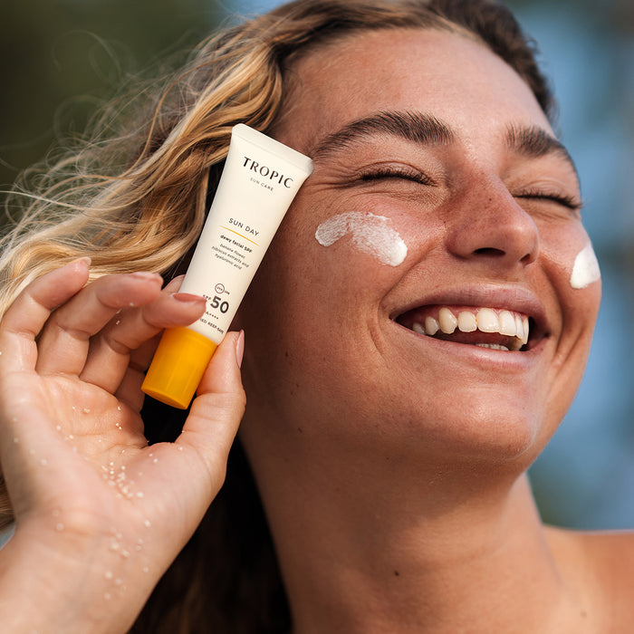 A smiling woman with sun-kissed skin and beachy waves holds a tube of Tropic Sun Care "Sun Day dewy facial  SPF 50 sunscreen" close to her face. She has two visible swipes of sunscreen on her cheeks and appears to be enjoying a sunny day outdoors, possibly at the beach, with sand on her hand and a blurred natural background.