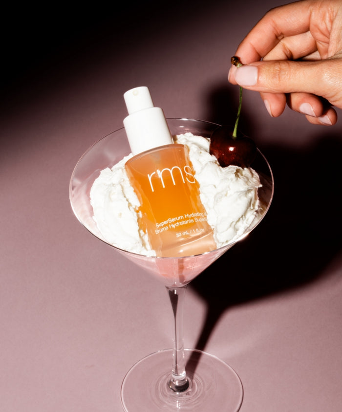 Bottle of SuperSerum Hydrating Mist nestled in whipped cream inside a clear glass, with a hand holding a cherry above it, highlighting clean beauty and natural ingredients.