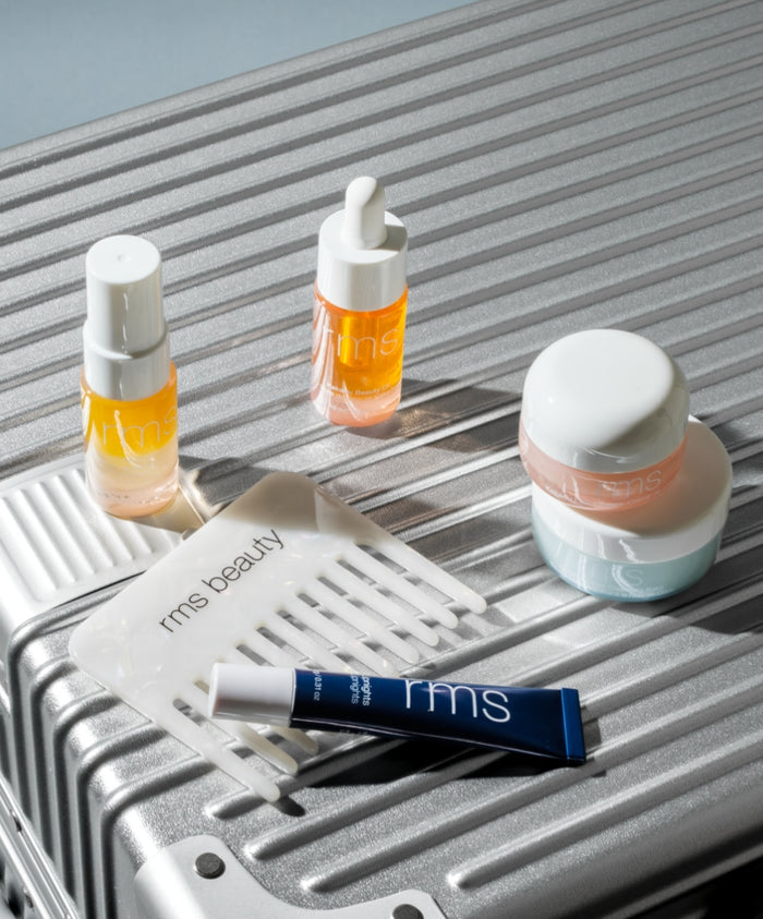 The Travel Skincare Set displayed on a silver textured surface includes small skincare bottles, jars, a hair pick, and a tube, showcasing clean beauty and sustainable packaging from RMS Beauty.